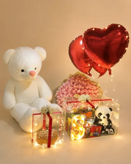Birthday surprise with large white teddy bear, cake in acrylic box, pink flower bouquet, snack hamper with panda teddy, red heart balloons and fairy lights