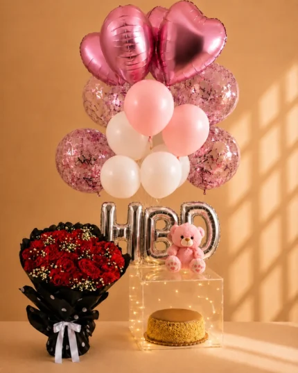 Birthday surprise setup with pink and white balloons, red rose bouquet, cake in illuminated acrylic box, pink teddy bear and silver HBD balloons