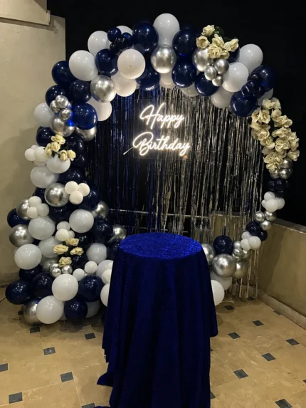 Navy blue white and silver birthday decoration with balloon arch, floral accents, metallic fringe backdrop and Happy Birthday neon sign in Lahore