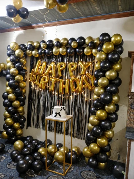 Black and gold balloon birthday decoration with Happy Birthday foil letters, fringe backdrop and cake stand in Lahore