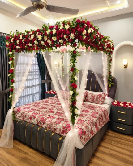 A denser red-and-white canopy crown with hanging floral accents and white draping framing the bed in a Lahore bridal bedroom