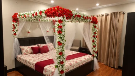 Artificial red-and-white canopy flowers with a lighter floral arrangement around the upper frame and white fabric below in a Lahore bridal bedroom
